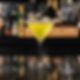 A vibrant yellow cocktail in a martini glass with a sugar rim sits on a reflective bar counter. Bottles are blurred in the background.