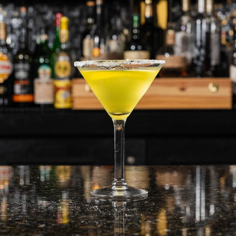 A vibrant yellow cocktail in a martini glass with a sugar rim sits on a reflective bar counter. Bottles are blurred in the background.