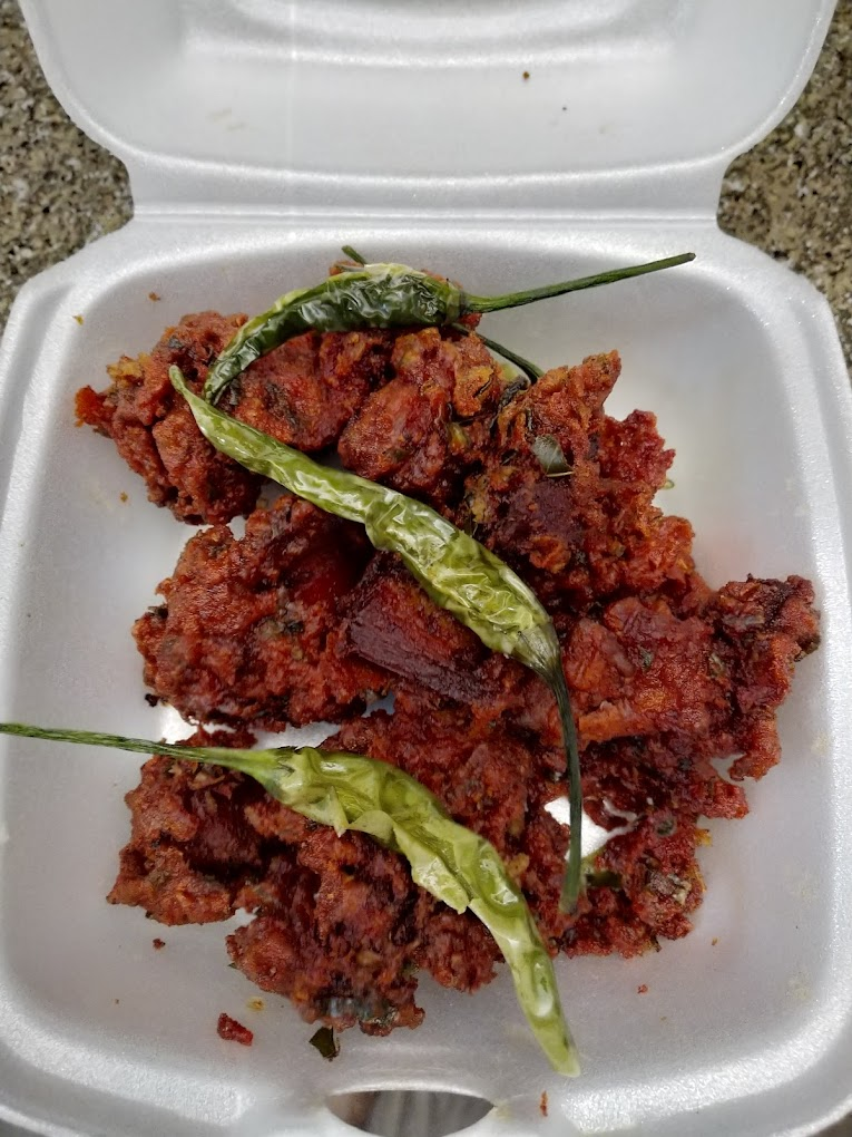 Spicy red curry with green chilies in a white foam container on a granite surface, creating a vibrant, appetizing look. No text visible.