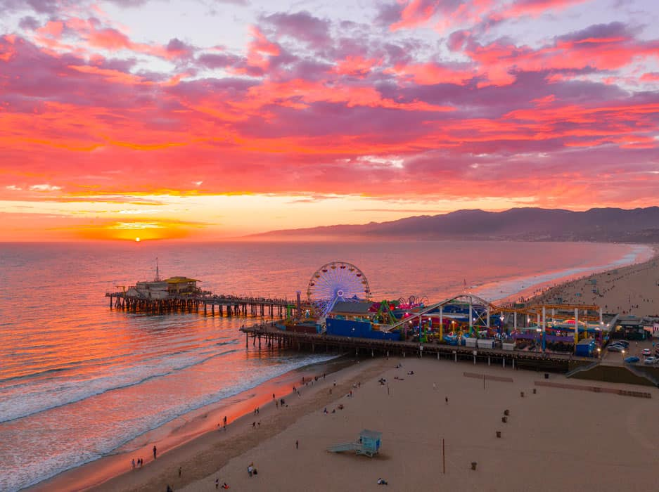 Beach at sunset with vibrant pink and orange clouds. A pier with a lit Ferris wheel stretches over the ocean. People are on the sandy shore.