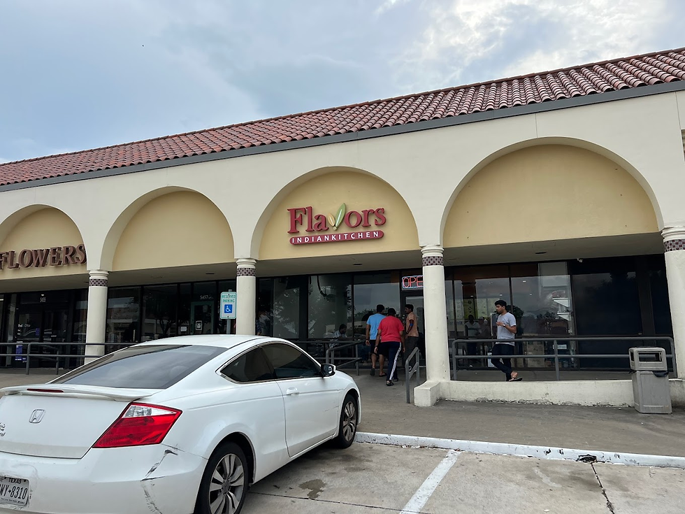Storefront of "Flavors Indian Kitchen" with people entering. A white car is parked outside. Overcast sky and "OPEN" sign visible.