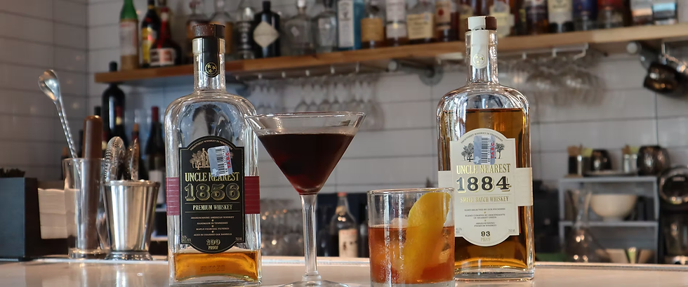 Whiskey bottles labeled "Uncle Nearest 1856" and "1884" on a bar with cocktails. Shelving with various bottles in the background.