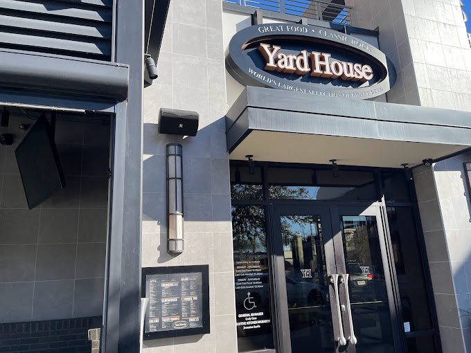 Yard House restaurant entrance with gray facade, large oval sign, and glass doors. Sunlight creates reflections; menu displayed outside.
