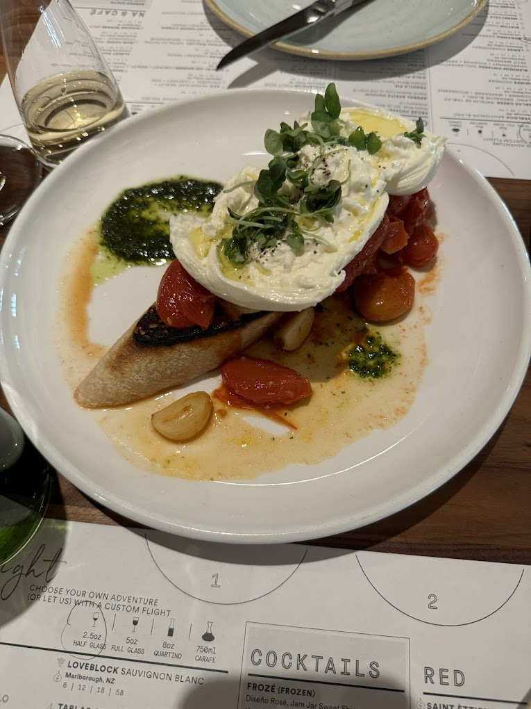 Toast topped with burrata, cherry tomatoes, greens, and pesto on a white plate. Wine glass and menu visible. Cozy dining atmosphere.