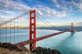 Red suspension bridge spanning blue water, city skyline in distance. Vibrant sky with clouds creates a serene, expansive atmosphere.