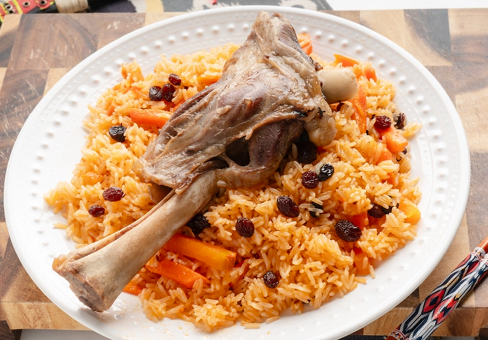 A plate of rice with raisins, carrots, and a lamb shank on a wooden table. Vibrant colors and patterned utensil evoke a warm, inviting feel.