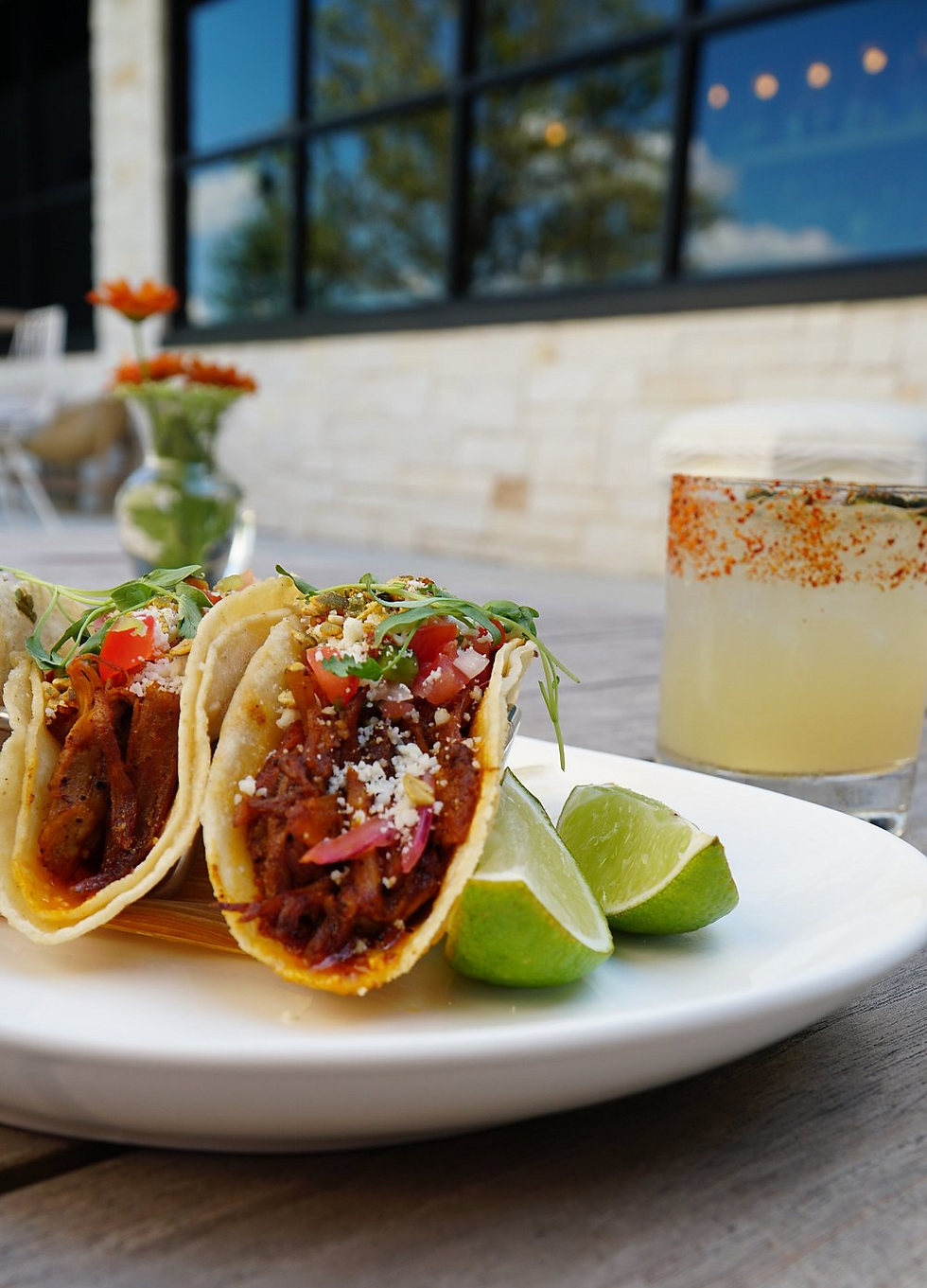 Tacos on a white plate with lime wedges, accompanied by a cocktail. Bright outdoor setting with flowers and brick wall in the background.