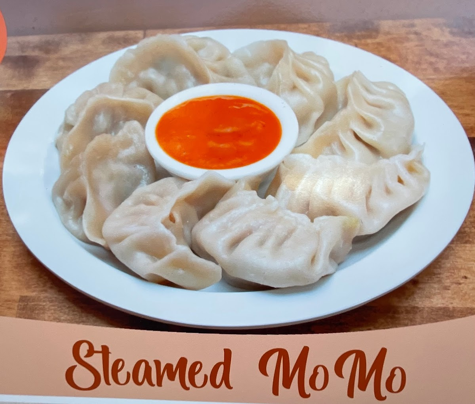 Plate of steamed momos with orange sauce in center on wooden table. "Steamed MoMo" text below in brown, casual font.