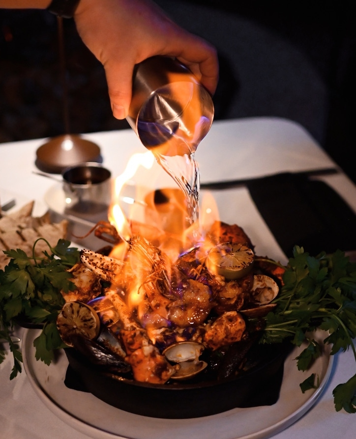 Hand pouring liquid from a metal cup onto flaming seafood platter with clams and shrimp. Parsley and lemon halves garnish the dish.