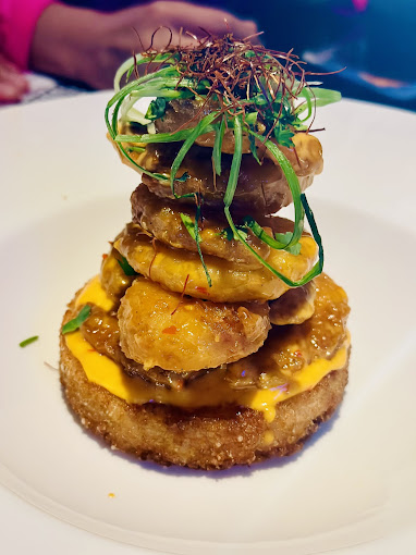 Stacked plantains and green garnish on a round breaded base with sauce. Warm lighting, restaurant setting. Vibrant and appetizing dish.