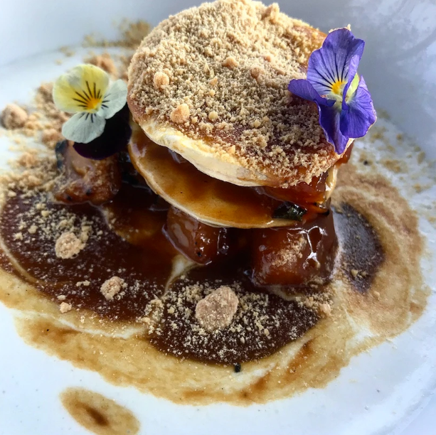 Gourmet dessert with layers of pastry, caramel sauce, and crumb topping, decorated with purple and yellow edible flowers on a white plate.