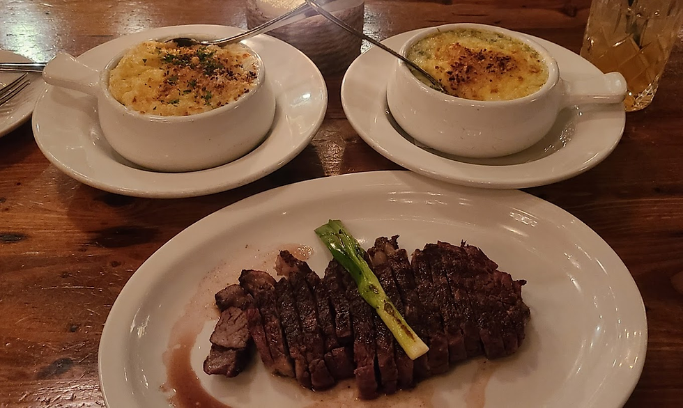 Steak with green onion on a white plate, two bowls of gratin, and a drink on a wooden table in a cozy setting.