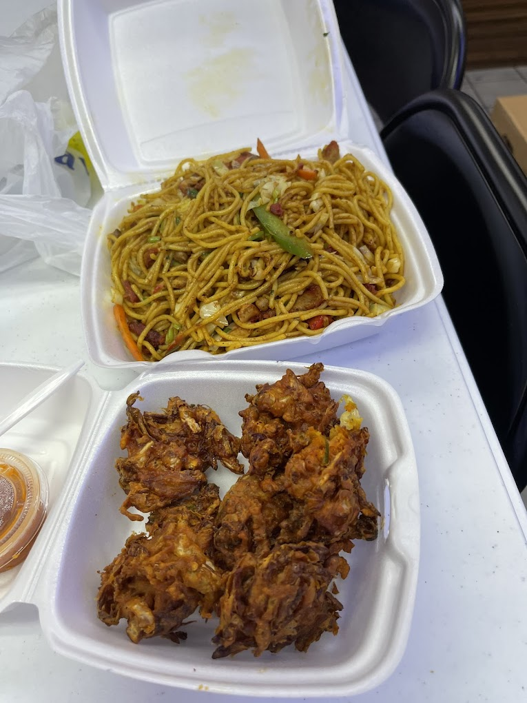 Takeout containers on a table: one with noodles and green peppers, the other with fried snacks. Sauce in a small cup nearby. Bright setting.