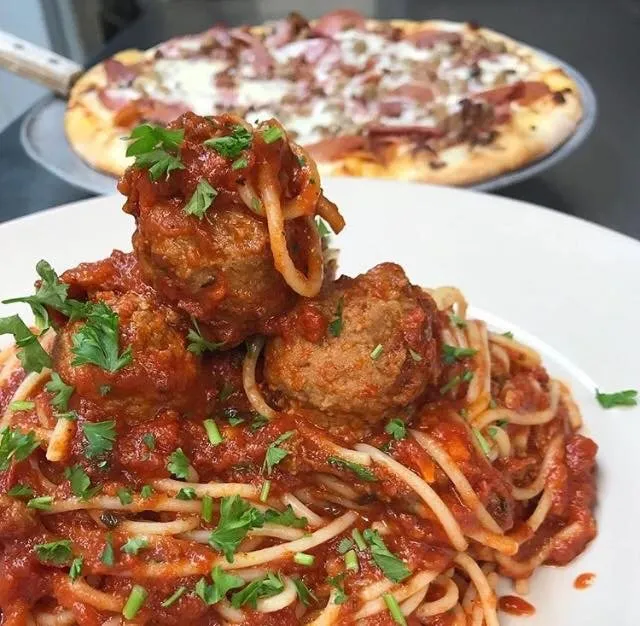 Spaghetti with meatballs in tomato sauce, garnished with parsley, on a white plate. A pizza is in the background. Cozy dining vibe.