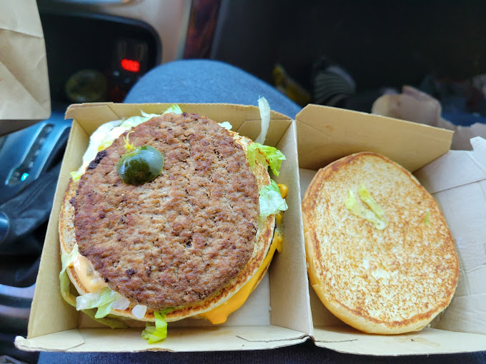 Burger with patty, lettuce, and sauce in a box, half-opened bun. Car seat background. Brown and green tones. Casual setting.