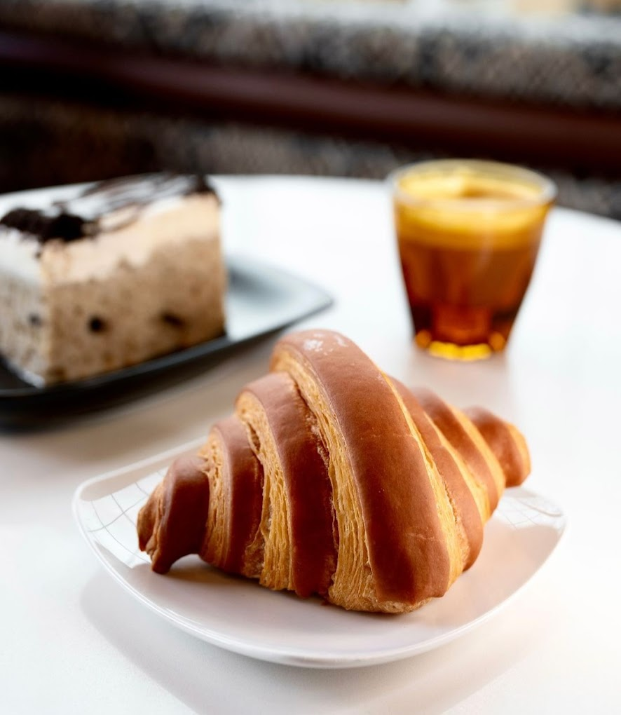 Croissant on a white plate with a slice of cake and an amber drink in the background on a table. Cozy, inviting atmosphere.