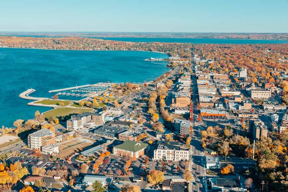 Aerial view of a lakeside city in autumn, showcasing vibrant fall foliage, buildings, and a marina by the blue water under a clear sky.