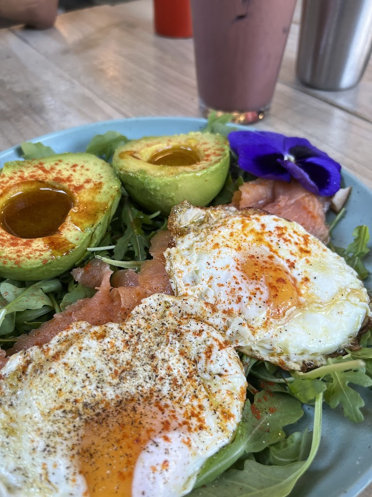 Breakfast plate with fried eggs, avocado, smoked salmon, arugula, and a purple flower. Pink smoothie and a silver tumbler in the background.