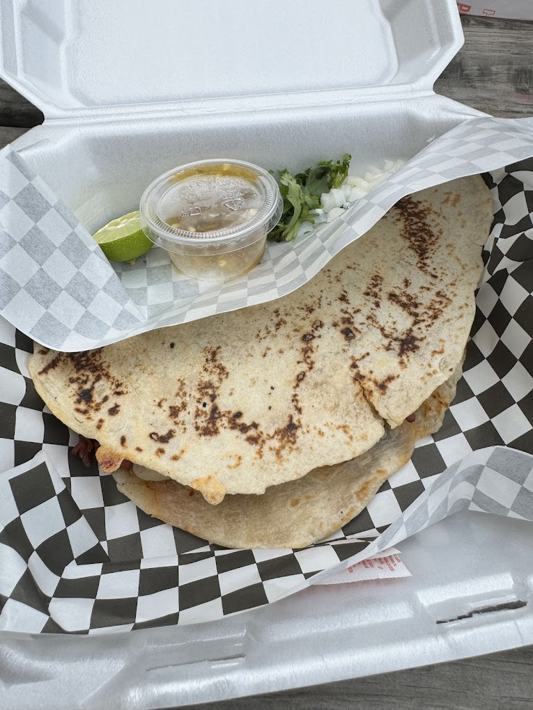Grilled quesadilla in a checkered paper-lined takeout box, with lime, herbs, onions, and salsa cup on the side. Brown and white hues.