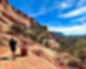 Person hiking on a red rocky trail with a wooden signpost and scenic mountain landscape. Clear blue sky, mid-day light.