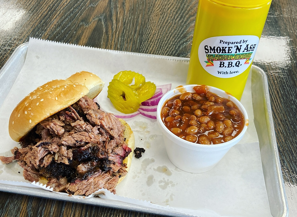 BBQ brisket sandwich with baked beans, pickles, and onions on a tray. Yellow BBQ sauce bottle labeled Smoke 'N Ash. Casual setting.