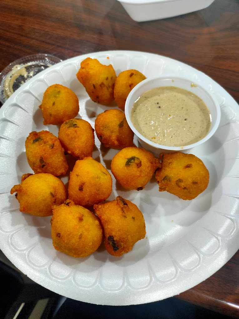 Fried snacks on a white plate with a cup of beige dip in a brown wooden setting. Snacks are round, golden-brown, with visible herbs.
