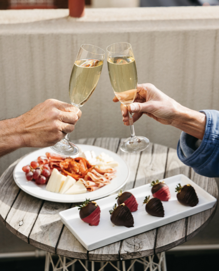 Two people toast with champagne flutes over a round table with a charcuterie plate and chocolate-covered strawberries. Cozy, celebratory mood.