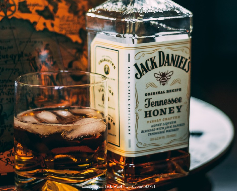 Close-up of a Jack Daniel's Tennessee Honey bottle next to a glass of whiskey with ice. Warm lighting and map in the background.