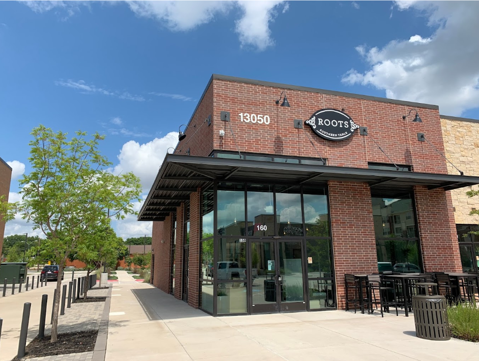 Brick restaurant with "ROOTS Southern Table" sign, number 13050. Sunny day, patio seating, and green trees. Blue sky with clouds.