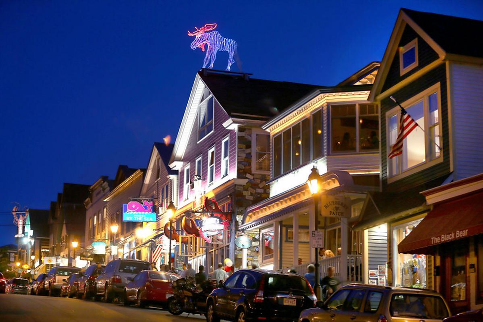 Colorful street at dusk with parked cars, lit signs, and a neon moose on a roof. People walk past shops with a festive, lively vibe.