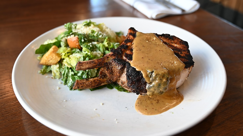 Grilled pork chop with gravy and a Caesar salad on a white plate, set on a wooden table. The dish looks savory and inviting.