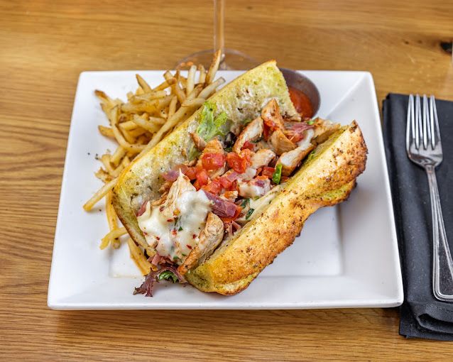 Gourmet sandwich with grilled chicken, melted cheese, and veggies on a white plate. Served with fries and sauce. Fork and napkin beside.