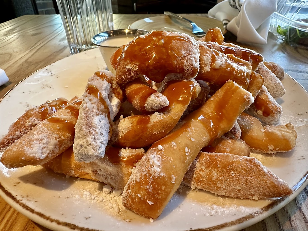 Plate of powdered sugar-coated churros drizzled with caramel on a wooden table. Glass and cloth napkin in the background.
