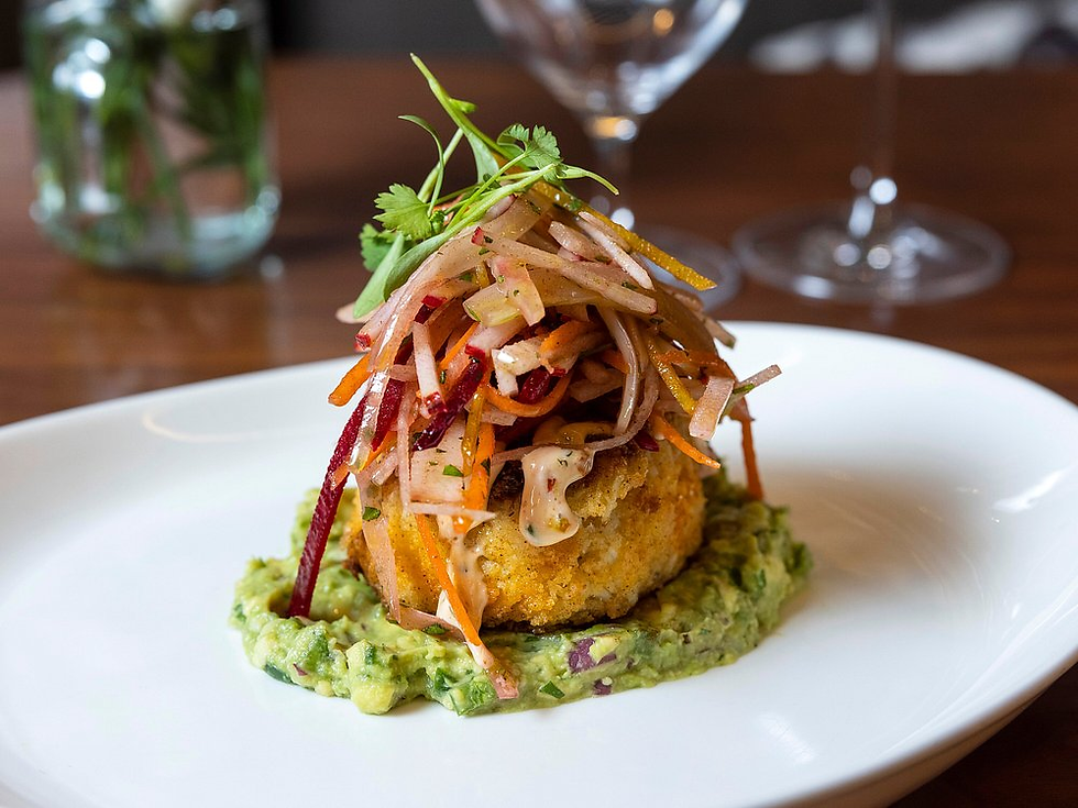 Gourmet dish on a white plate: a fried cake topped with julienned vegetables sits on guacamole. Background features blurred glasses.
