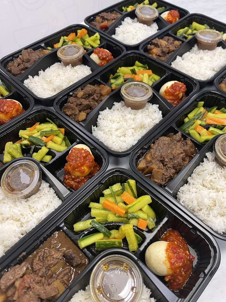 Black meal trays with white rice, beef stew, boiled eggs, and mixed vegetables. Sauces in small containers. Bright colors and appetizing display.
