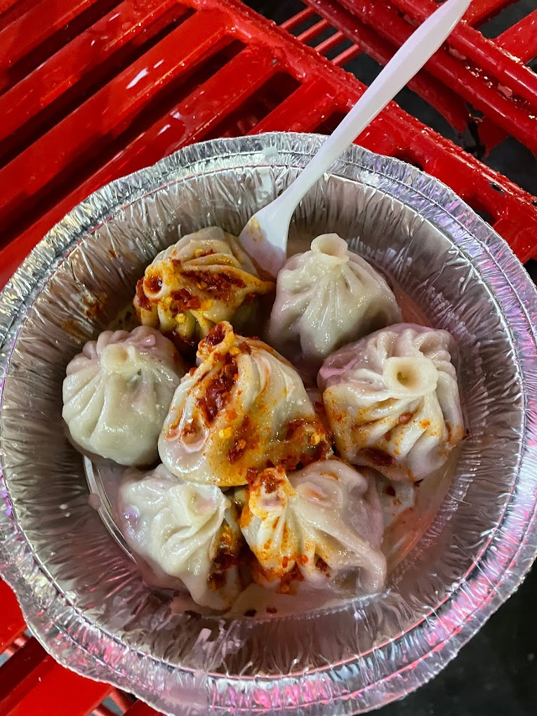 Six dumplings topped with spicy sauce in a foil bowl, resting on a red plastic table. A white plastic spoon is placed in the bowl.
