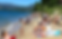 Beach scene with people sunbathing and playing by the water. Clear blue sky, green trees, and mountains in the background. Relaxed mood.