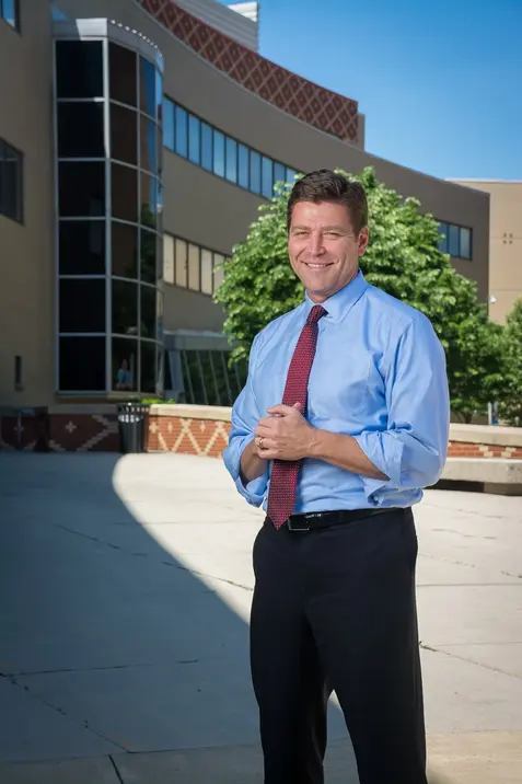 Professional outdoor portrait of a person in a light blue dress shirt and tie standing in front of a modern building with large windows.