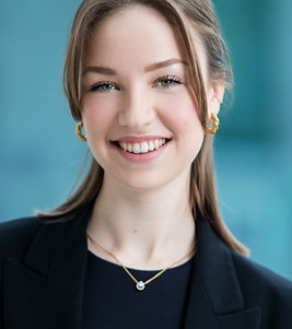 Das Bild zeigt Franzi. Eine junge Frau mit mittellangen blonden Haaren, die sanft nach hinten gelegt sind. Sie steht vor einem unscharfen, blau-türkisen Hintergrund, trägt goldene Ohrringe, eine feine Halskette und ein dunkelblaues Oberteil mit schwarzem Blazer. Sie lächelt freundlich in die Kamera.