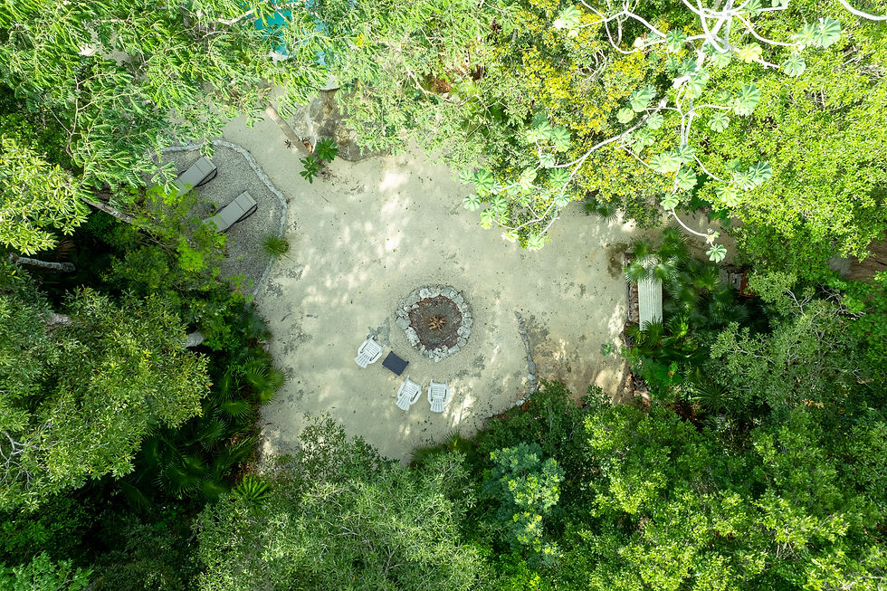 Aerial view of a sandy area with three white lounge chairs around a fire pit, surrounded by lush green trees and a small bench. Relaxing vibe.