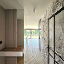 Jasny korytarz z tapetą w liście palmowe, lustrem, drewnianym siedziskiem i podłogą w jodełkę, prowadzący do ogrodu.