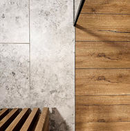 Kontrastowe połączenie w łazience: szerokie deski drewnopodobne obok szarych płytek ceramicznych (imitacja kamienia/lastryko), detal drewnianej ławki.