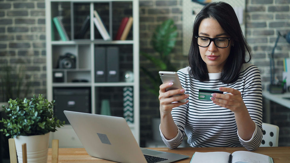 Photo of a dark-haired woman in a striped top wearing dark rimmed glasses looking at her phone and credit card while having her laptop open, suggesting that she's shopping online. An attractive green plant sits to her right as a decoration. The backgrounds is a brick wall with a neat bookcase. Photo by Vitaly Gariev on Unsplash