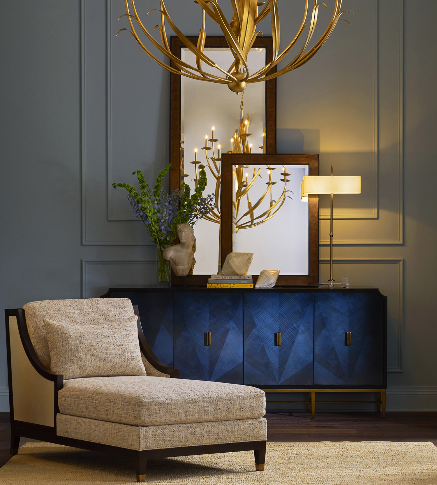 Elegant living space with a beige chaise lounge, navy blue sideboard, layered mirrors, brass table lamp, and gold branch chandelier against gray paneled walls
