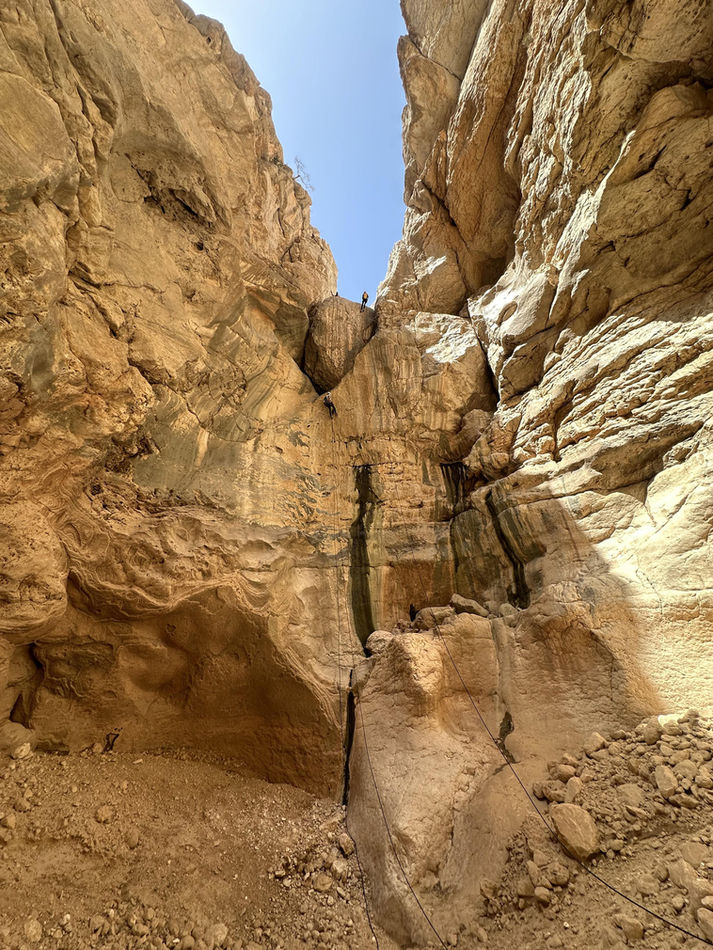 Narrow sandstone canyon walls on the Wadi Ta’ab adventure route in Oman
8 Water-filled canyon corridor in Wadi Ta’ab during a guided canyoning experience in Oman
9 Waterfall and lush canyon section on the Wadi Ta’ab canyoning trail in Oman
10 Hikers ascending a rocky slope on the Wadi Ta’ab canyoning approach in Oman
11 Canyoner walking through a narrow gorge during the Wadi Ta’ab one-day adventure tour
12 Traveler standing in a canyon pool beneath mossy rock walls in Wadi Ta’ab, Oman
13 Participants abseiling into a rocky canyon pool during the Wadi Ta’ab canyoning tour in Oman
14 Canyoner rappelling beside a waterfall on the Wadi Ta’ab canyoning adventure in Oman
