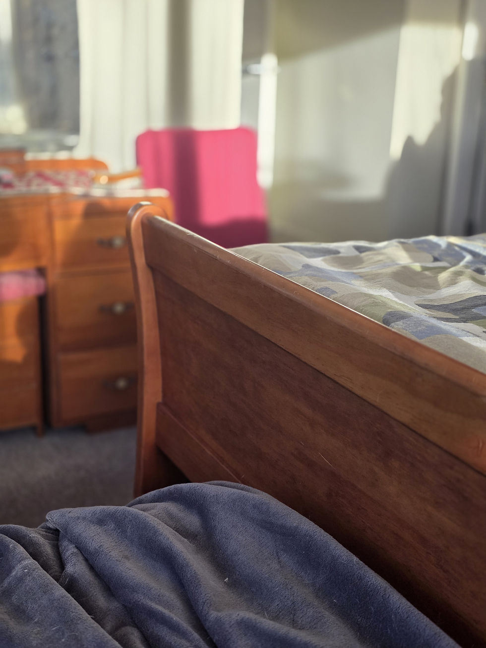 Sunlit bedroom with a wooden bed, blue blanket, dresser, and magenta coloured chair.