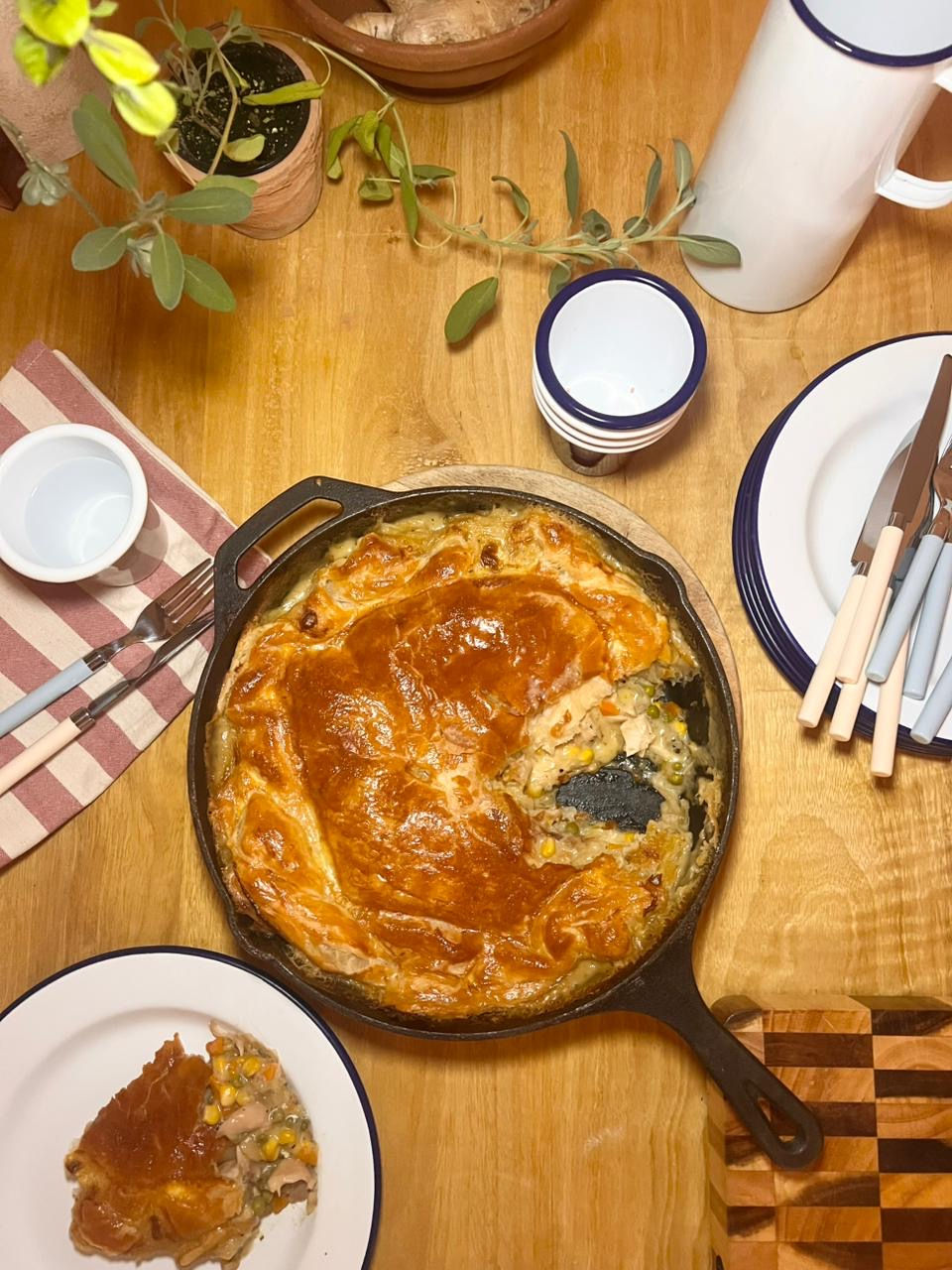 Rustic dining setting with a golden pie in a skillet on a wooden table, surrounded by plants, cups, and a red-striped napkin.