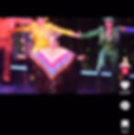 Performer in a sparkly multicolored outfit and headdress dances on stage with colorful backup dancers in yellow and green outfits.