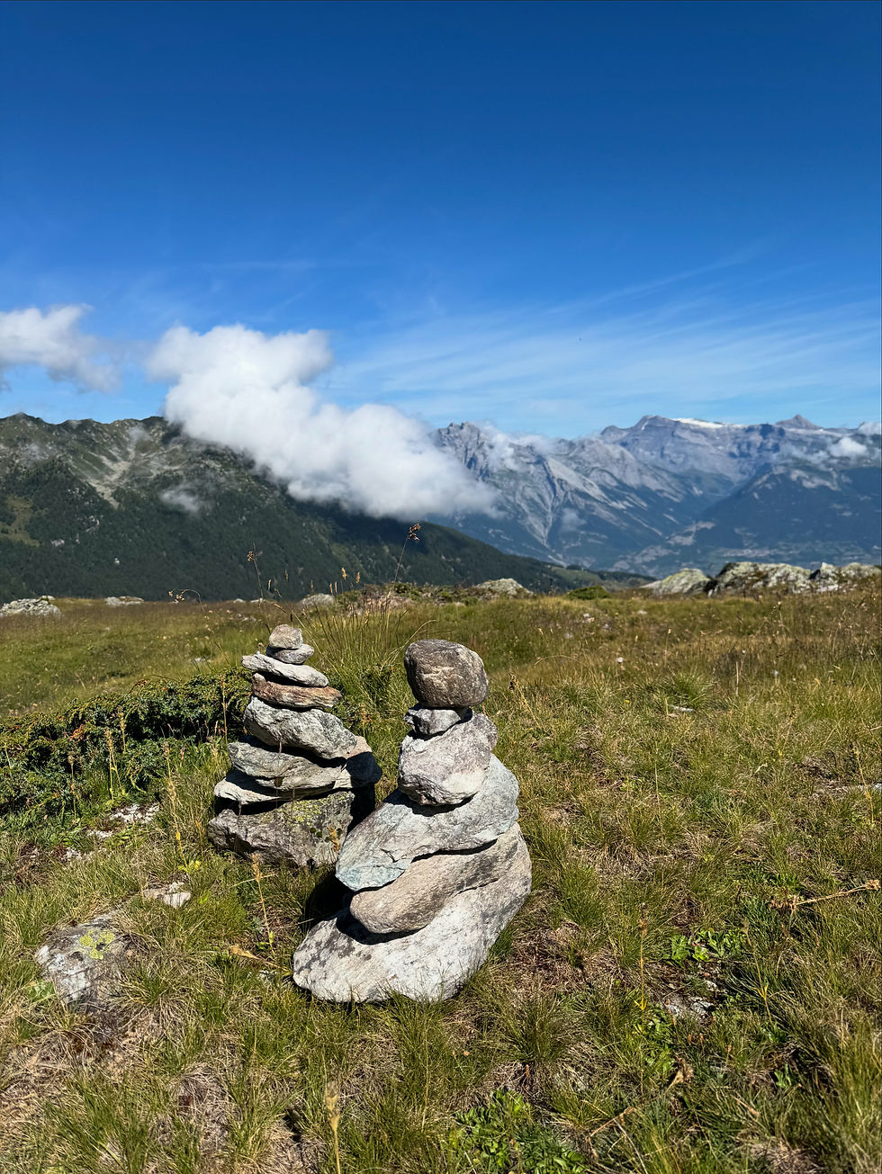 Steenmannetje langs een bergpad, met uitzicht op een vallei, alpenweide en veel lucht, symbool voor stilstaan, richting en innerlijke rust.