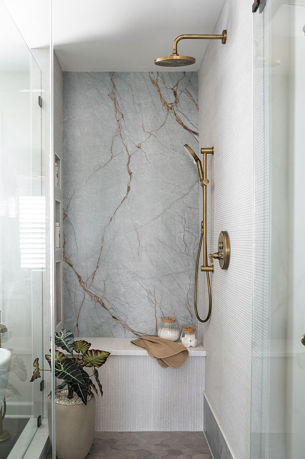 Sleek bathroom with a marble shower, brass fixtures, potted plant, and beige towel. Glass partition and muted color tones create a serene vibe. marble slab, marble bathroom, porcelain slab, gold bathroom fixtures, gold faucet, gray bathroom tile